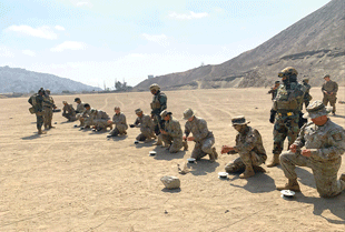 A group of people in beige military uniforms kneel in a row on sandy ground and work with small rolls of wire. 