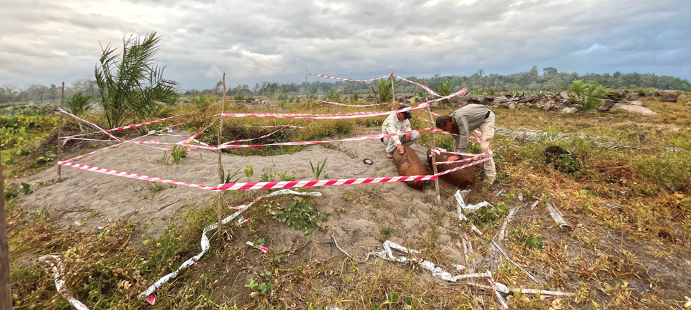 Two men crouch around rusty bombs in a roped of area.