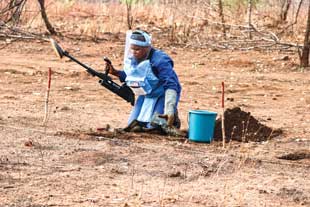 A person kneeling in a field holding a metal detector while searching for landmines.