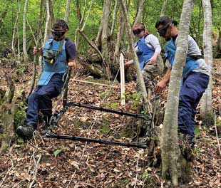 Three people in protective hear carry a large loop slightly above the ground through wooded land