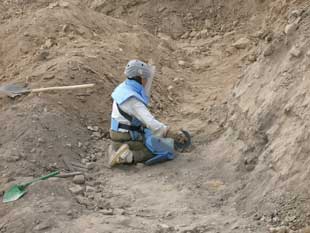 A person in protective gear kneeling in a large dirt pit looking for explosive hazards.