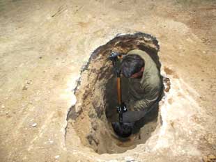 A man with a shovel standing in a narrow dirt pit 