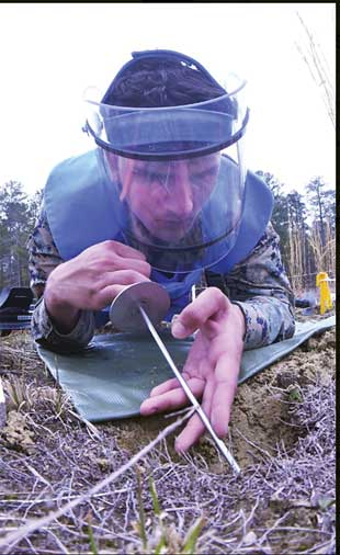 A person in protective gear laying on his stomach probing the ground with a metal stick