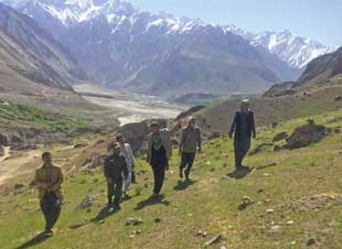Six people walk up a steep rocky incline with a valley and mountains in the background