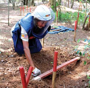 A kneeling person in protective gear with a tool in her hand digging in the dirt for a landmine