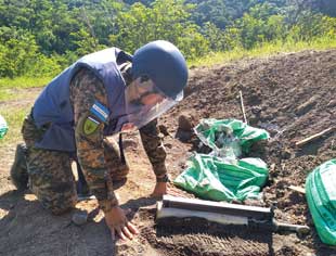 A man in protective gear kneeling in the dirt beside a metal object