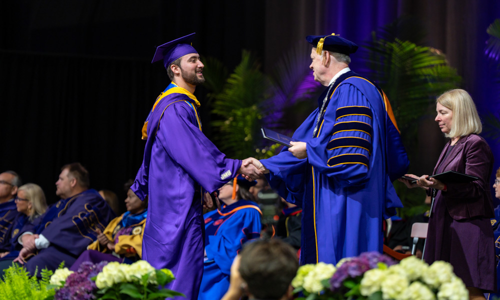 Graduate in a purple cap and gown shakes hands with a faculty member in blue regalia on a commencement stage.