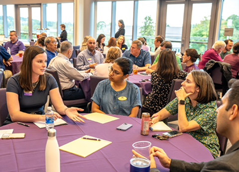 A group of attendees engaged in conversation at a networking event, seated around tables with purple tablecloths.
