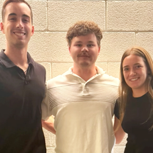 Three people standing side by side, smiling in front of a light brick wall.
