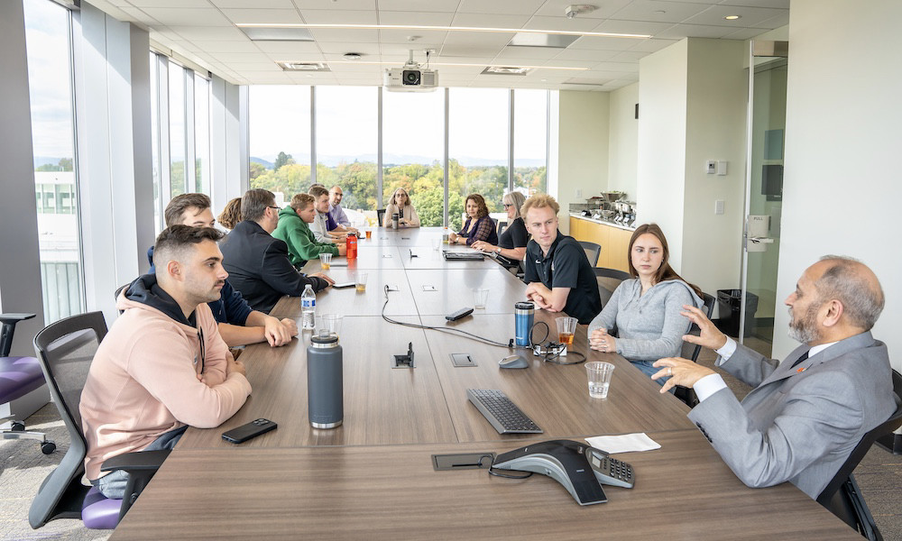 Several men and women sit around a large, oval conference table with floor to ceiling glass windows all around. 