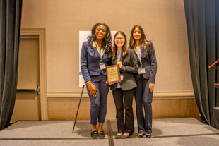 Three women standing on a stage at an event, holding a plaque and smiling.