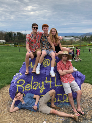Five people sit and lean on a large painted purple rock outdoors on a grassy field. The rock features yellow and blue lettering related to a Relay for Life event. The group is casually dressed in warm‑weather clothing, and a few other people can be seen walking in the background under a cloudy sky.