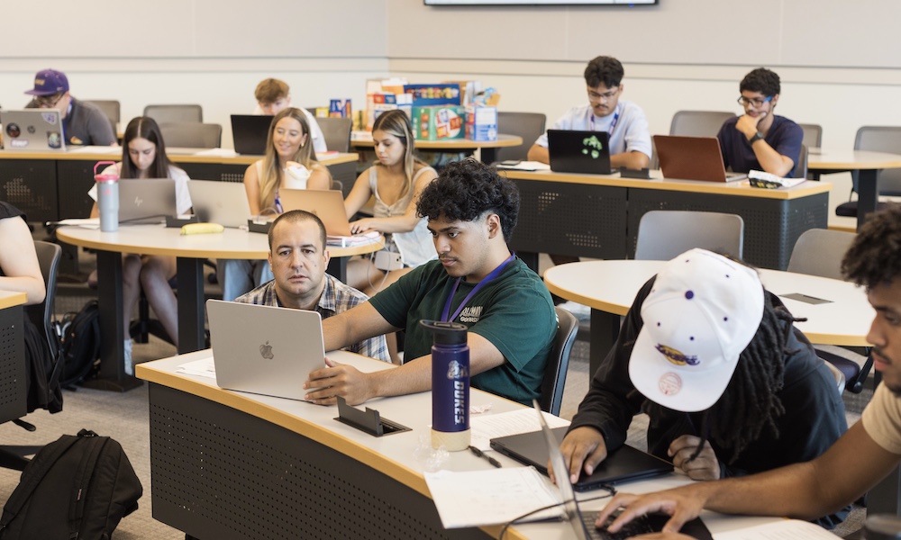 A classroom scene featuring students engaged in learning, some using laptops while others participate in discussions.