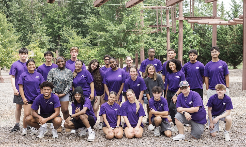 A group of diverse young people wearing purple shirts poses together outdoors in a park setting.