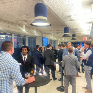 People networking at a corporate reception, standing around cocktail tables under hanging pendant lights in a bright office space.