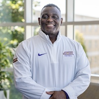 Charles T. May Jr. smiling in a white athletic quarter-zip jacket standing indoors near large windows.