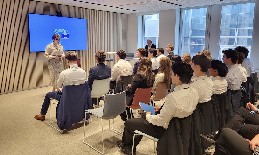 A presenter speaks to a group of seated young adults in a modern conference room with a European Union logo displayed on a screen.