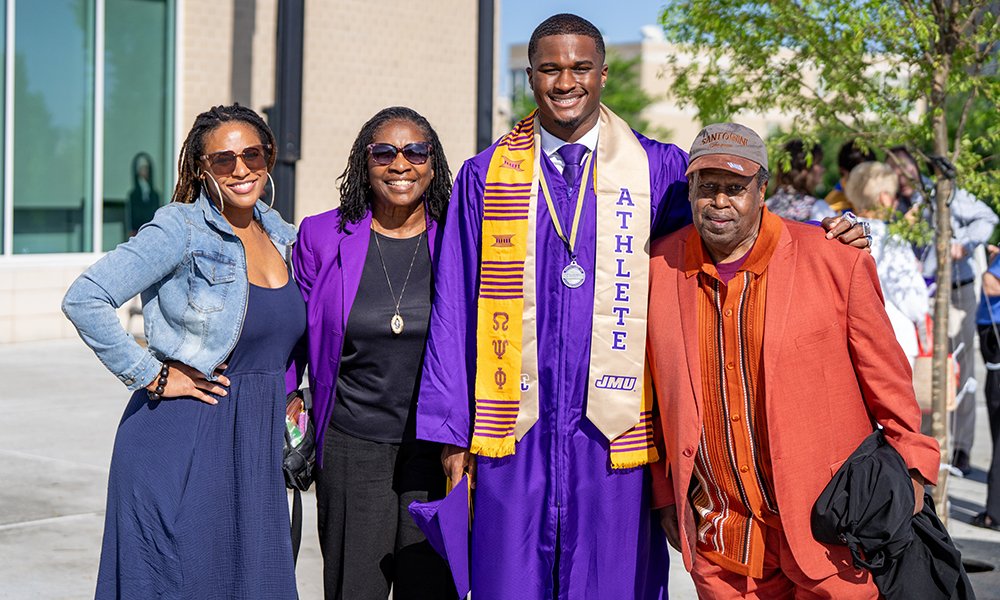 A smiling graduate in a purple cap and gown stands with three family members, celebrating his achievement at a graduation ceremony.