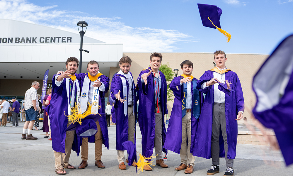 A group of six male graduates in purple gowns tossing their caps in celebration outside the Atlantic Union Bank Center.