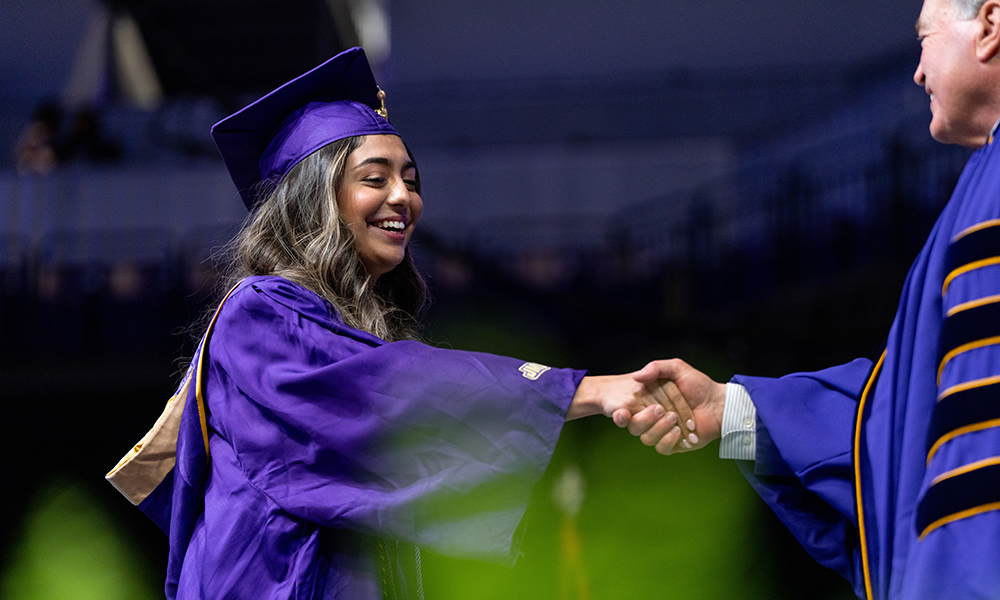 A graduate in a purple cap and gown happily shakes hands with a professor during a commencement ceremony.
