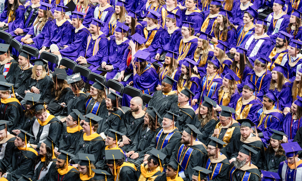 Large group of graduates seated at a commencement ceremony, wearing purple and black caps and gowns with gold stoles.