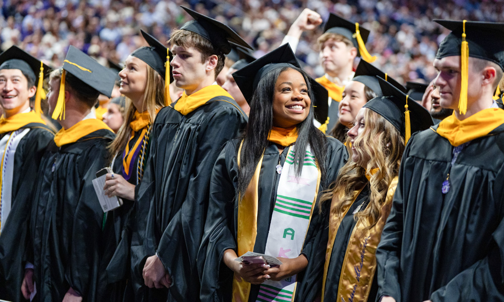 Group of graduates in black caps and gowns with gold stoles standing in a crowded ceremony, one smiling in the center.