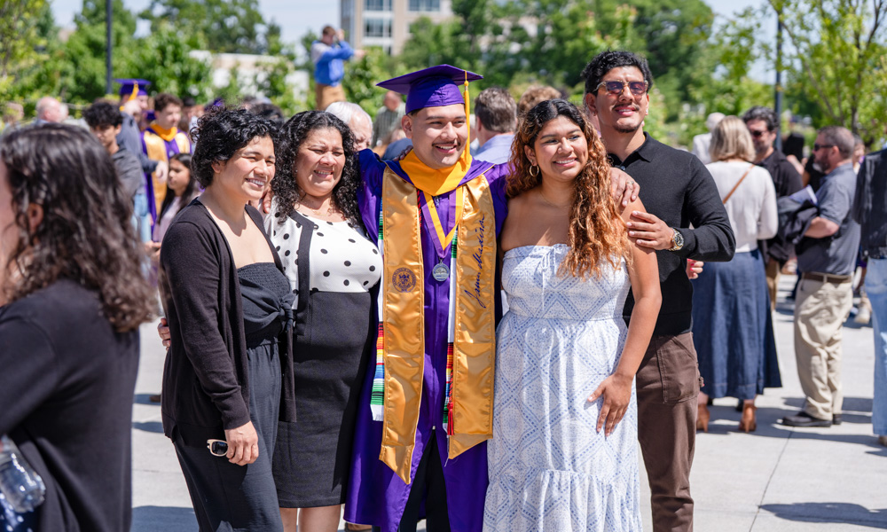 Graduate in a purple cap and gown poses with family outdoors at a graduation ceremony, smiling in a crowd.