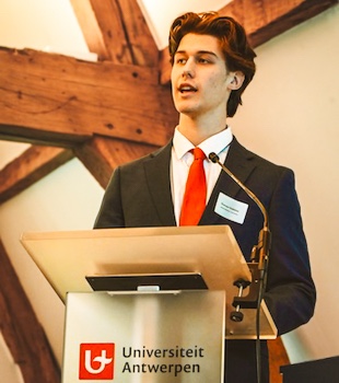 A young man in a suit delivers a speech at a podium featuring the Universiteit Antwerpen logo.