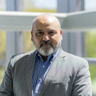 Elias Semaan with a beard, wearing a gray blazer and blue striped shirt, standing in a bright, modern environment.