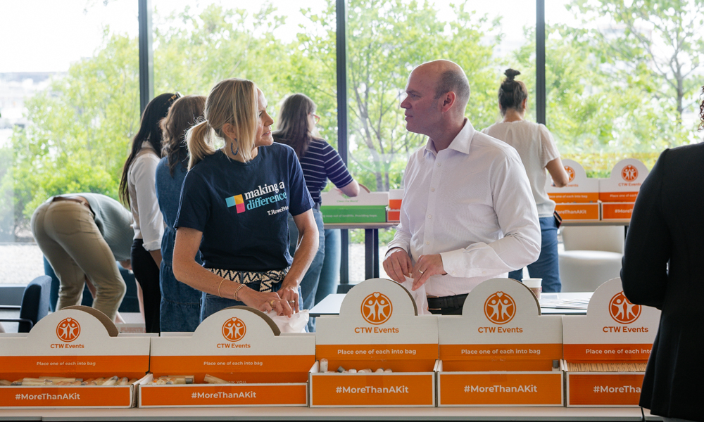 Volunteers pack charity kits at a table of orange-and-white CTW Events boxes labeled #MoreThanAKit, while two people talk in the foreground by large windows.