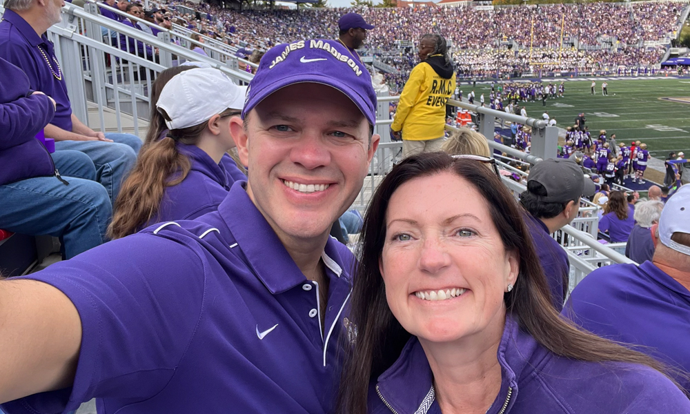 Eric Veiel and his wife Lori in purple take a smiling selfie in the stands at a crowded football stadium during a game.
