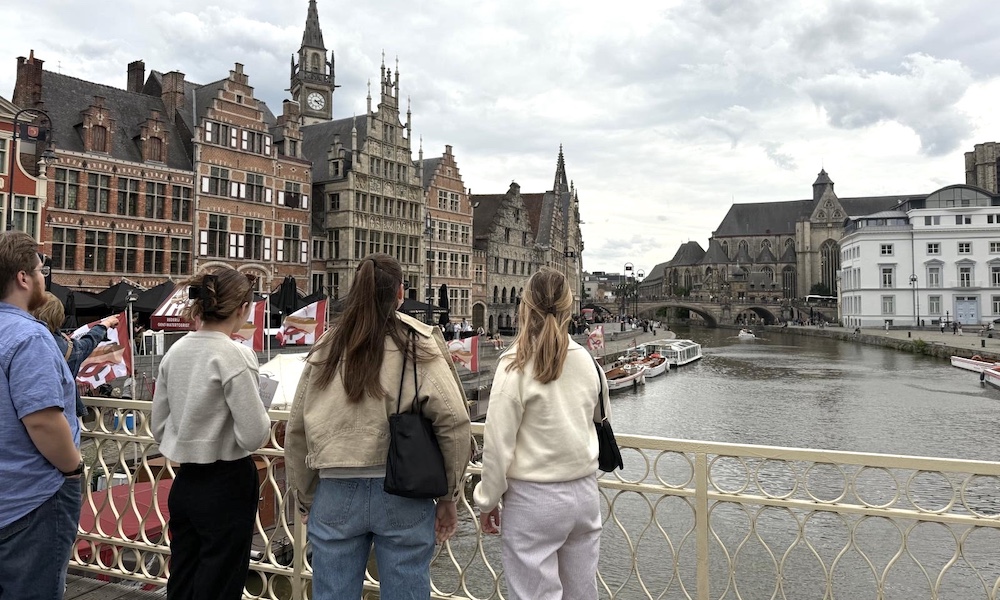 A group of students standing by the Leie River, gazing at historic buildings and boats in Ghent, Belgium.