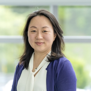 Headshot of Hui Sono with shoulder-length dark hair, wearing a white blouse and a purple cardigan, smiling warmly.