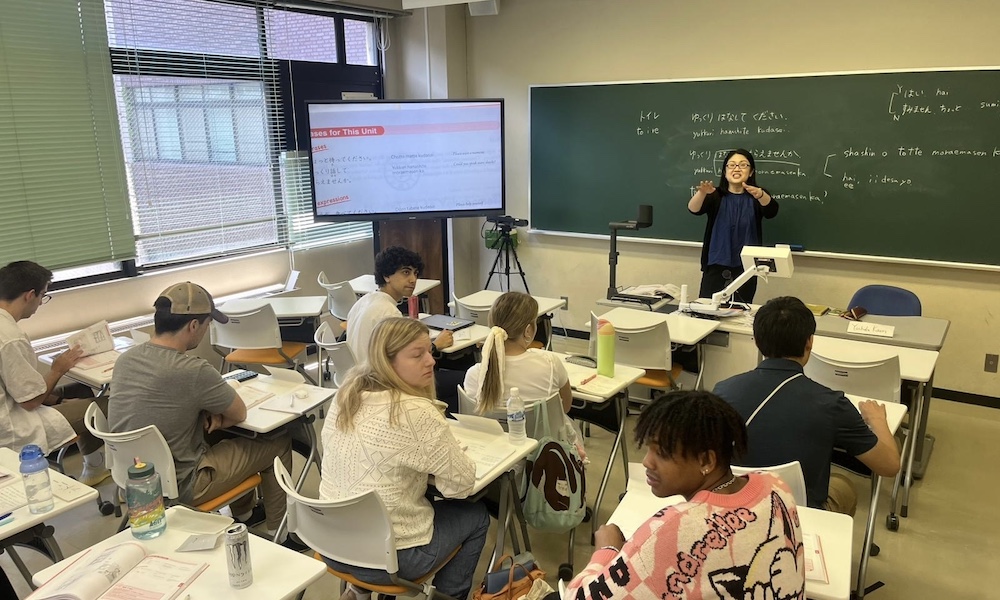 A classroom setting in Japan with a teacher instructing students; the students are engaged and taking notes while the teacher presents information on a board.