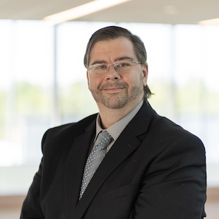 Professional headshot of a Jeremy Ezell in a suit and tie, smiling confidently in a modern office setting.