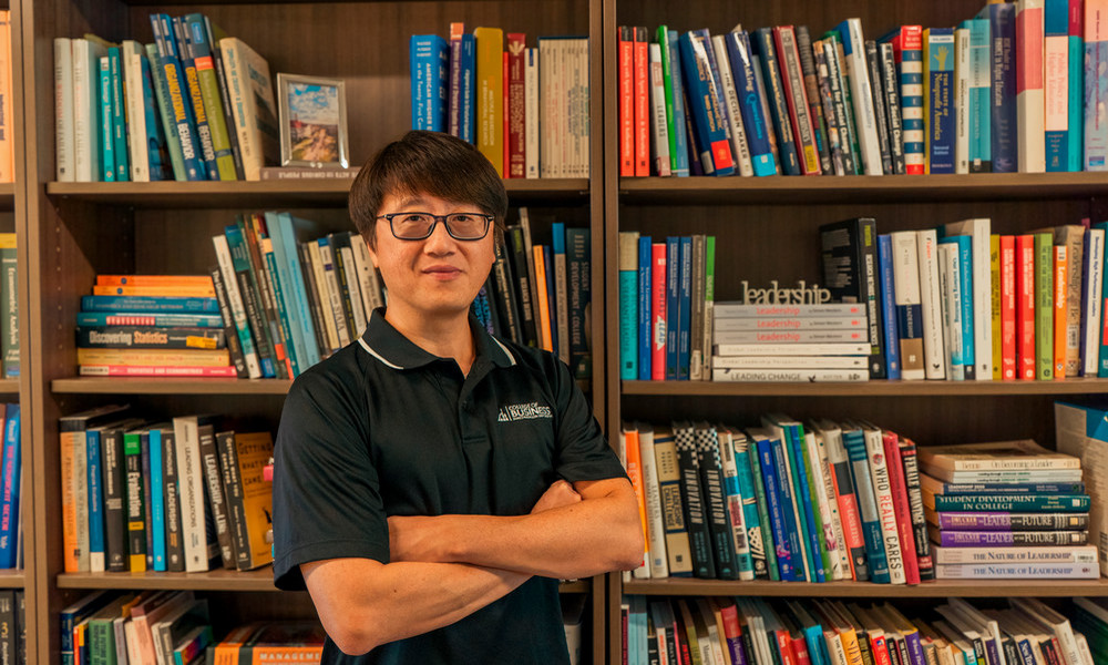Professor John Guo wearing a black collared shirt with a “College of Business” logo stands with arms crossed in front of two large bookshelves filled with academic and business-related books. Some visible titles include “Leadership” and “Discovering Statistics.”