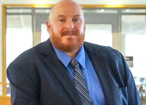 Portrait of John Lauck wearing a navy suit, blue shirt, and striped tie in an office setting.