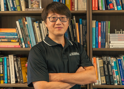 Dr. John Guo, professor of Computer Information Systems and Business Analytics, stands confidently with his arms crossed in front of a bookshelf filled with various books on leadership and management.