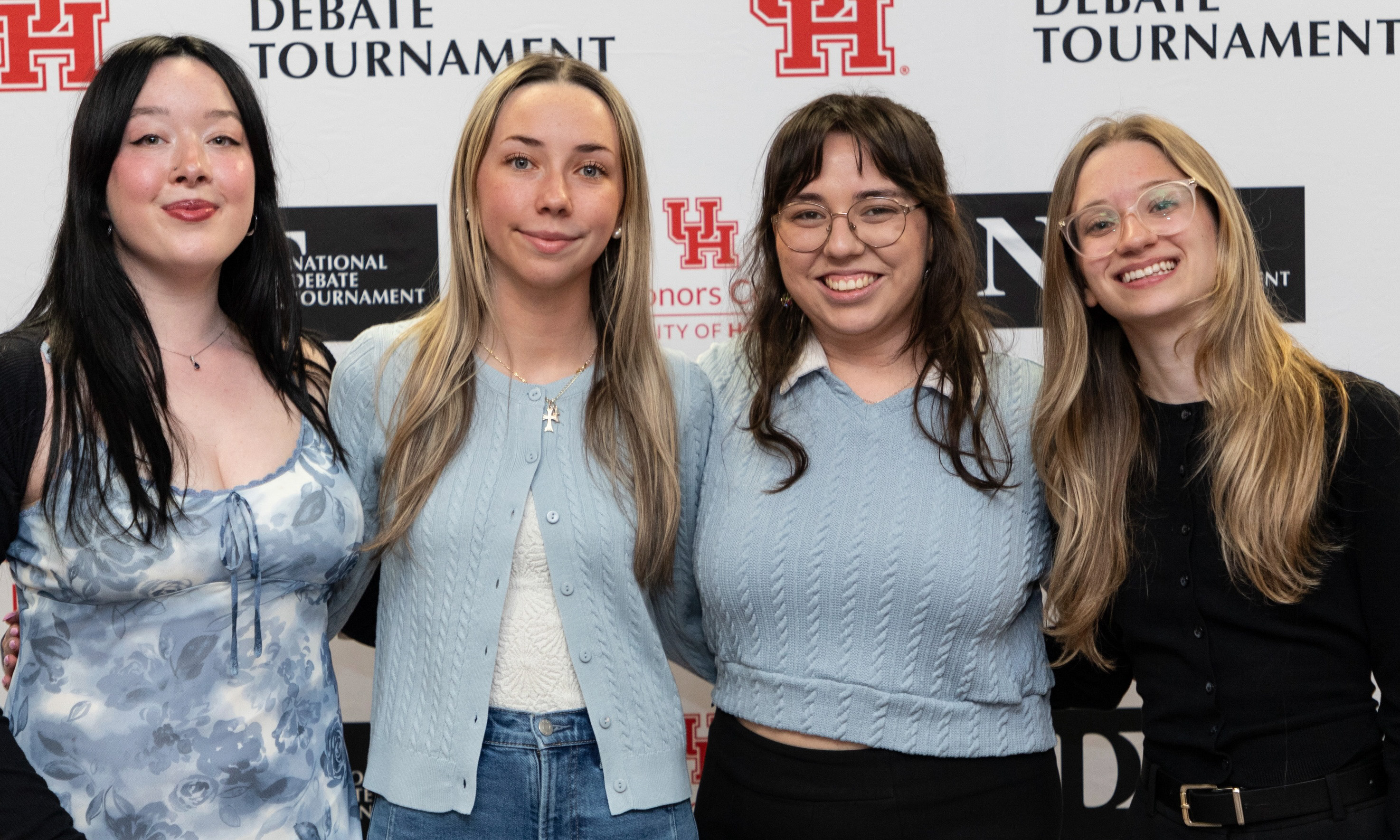 Four students pose and smile in front of a University of Houston debate tournament backdrop.