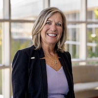 Smiling business professional in a black blazer standing indoors with large windows in the background.