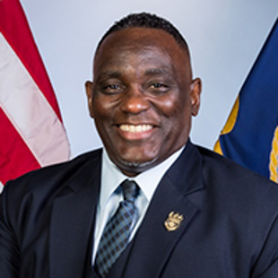 Headshot of Charles T. May Jr. in a suit, standing in front of American and state flags.
