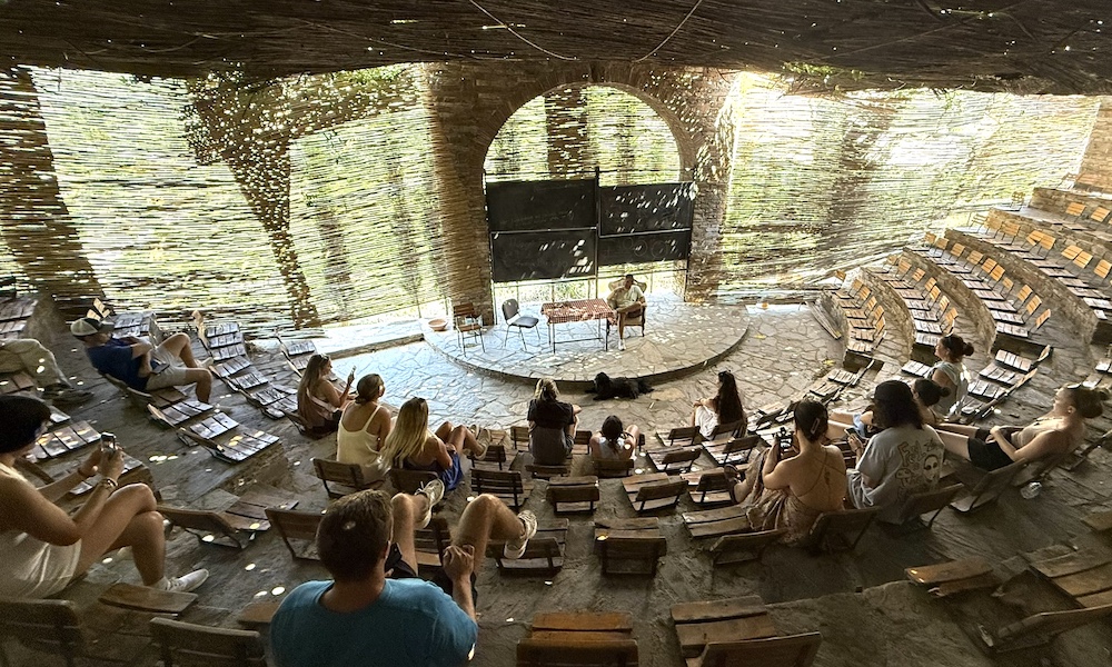 A rustic amphitheater filled with people seated on wooden benches, surrounded by natural elements and a woven canopy.