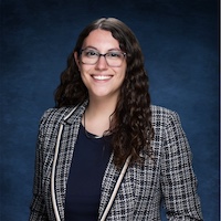 Smiling woman with long curly hair and glasses wearing a plaid blazer in a studio portrait.
