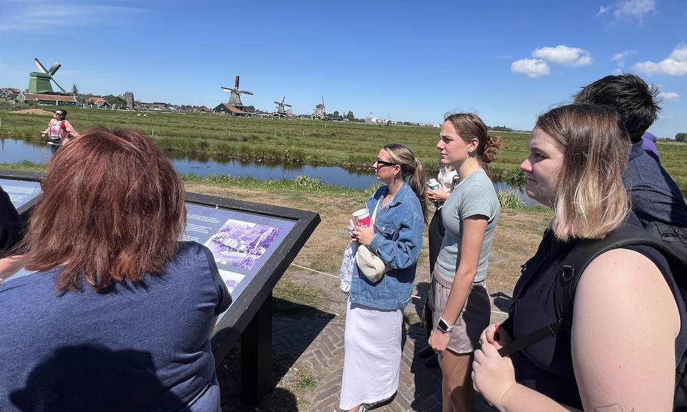 A group of people observing an informational display near a scenic landscape with windmills in the Netherlands.