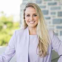 Smiling woman in a light lavender blazer standing outdoors near a brick wall.
