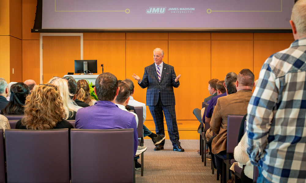 President Jim stands in the center of the room. with faculty and staff seating to the left and right of an aisle of chairs. He's talking with his hands and is wearing a plaid suit with a purple and gold tie. 