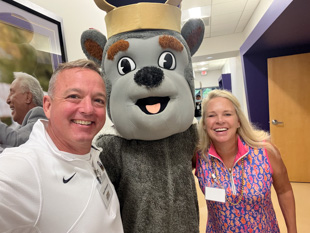 Dave and Alisa pose for a selfie with Duke Dog in a hallway.