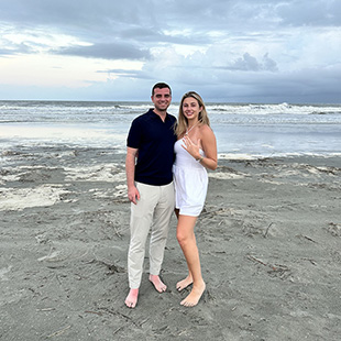 Tyler, on the left wearing a black shirt and khakis stands with Jordan who is wearing a white romper with her hand up showing off her new engagement ring., They are standing on the beach with the ocean behind them.  