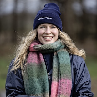 Taylor Holmes smiling, wearing a navy beanie and a colorful striped scarf, posing outdoors.