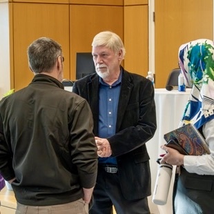 A small group of people stand in conversation in a bright, modern indoor space. One person holds a notebook and a rolled‑up poster, while another gestures with their hands. Wood paneling and a podium are visible in the background.